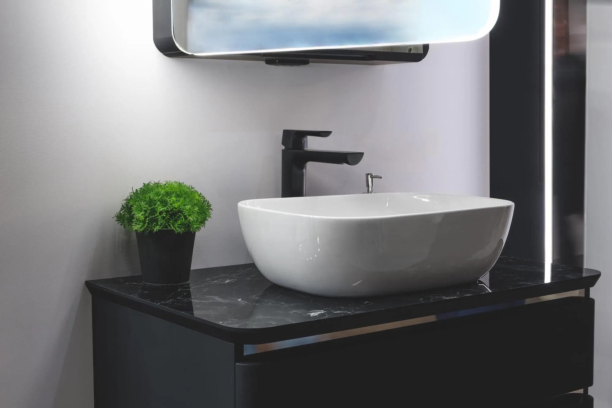 Modern minimalist bathroom interior with a white sink on a black marble countertop with a black tall faucet and a mirror with blue lighting and a black pot with a green decorative plant