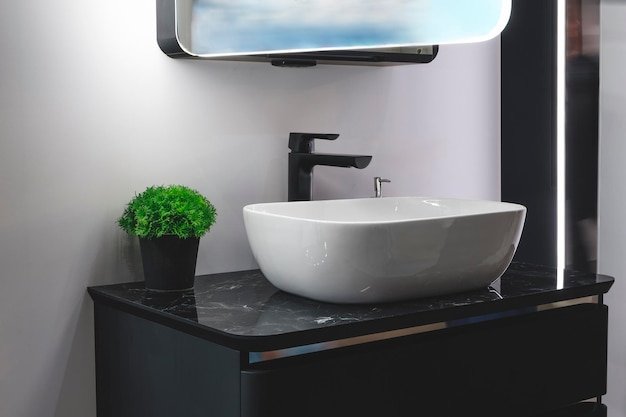 Modern minimalist bathroom interior with a white sink on a black marble countertop with a black tall faucet and a mirror with blue lighting and a black pot with a green decorative plant