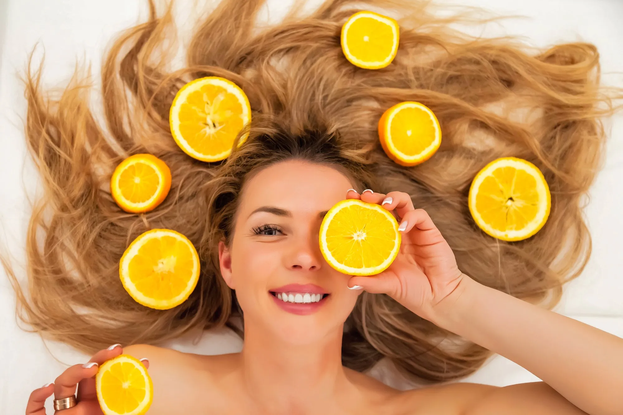 Pretty charming joyful attractive cheerful positive young woman with long hair having pieces of orange, close one eye on white background. Concept of beauty, health and personal body care. Copy space
