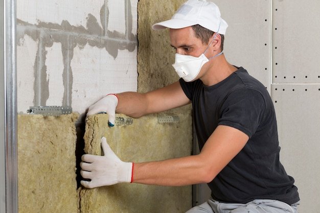 Worker in protective respirator mask insulating rock wool insulation in wooden frame for future house walls for cold barrier. Comfortable warm home, economy, construction and renovation concept