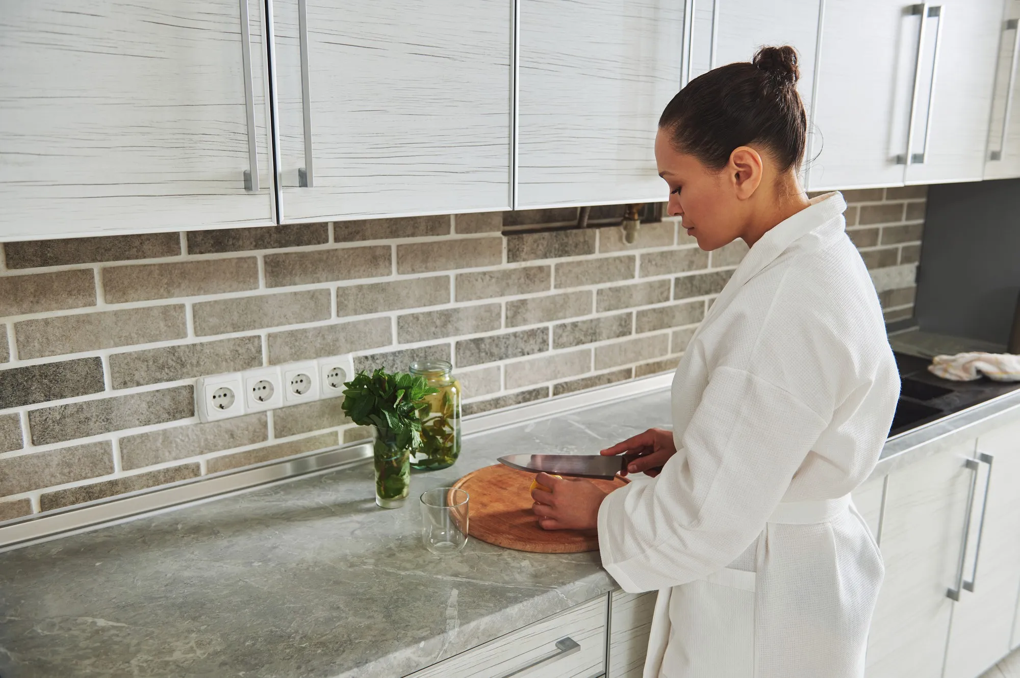 Young beautiful woman in white waffle bathrobe cutting lemon on a wooden board to prepare lemon mint water. Morning routine, healthy eating concepts