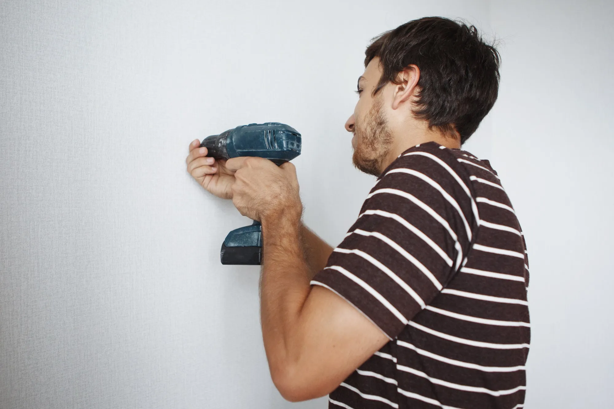 Young handsome guy with a screwdriver is drilling a white wall in a new apartment doing repairs.