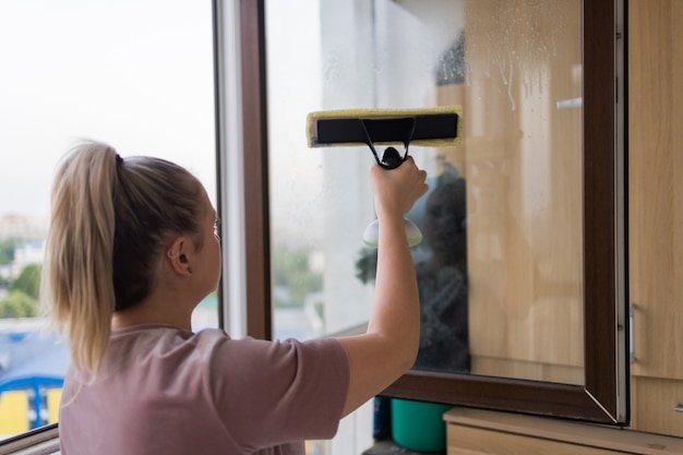Beautiful young housewife in working clothes washing windows by professional tools at home.