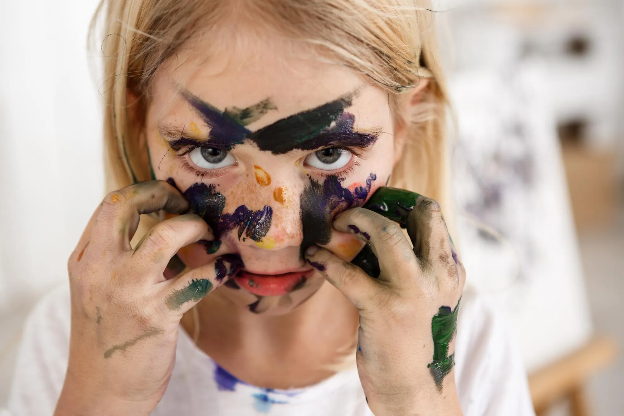 Close up portrait of beautiful little European girl with blond hair and freckles looking at the camera with her big eyes and touching her face with hands in paint