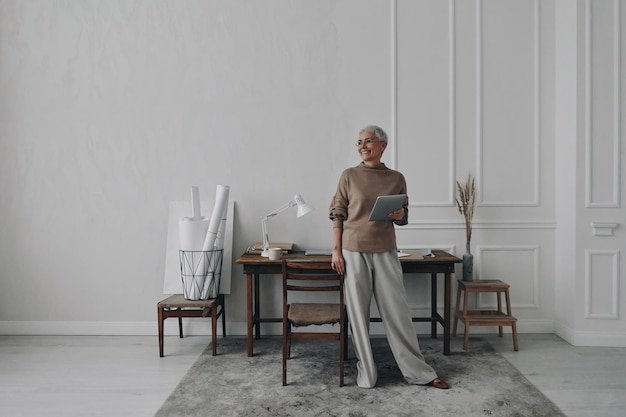 Happy senior woman holding digital tablet while standing near her working place