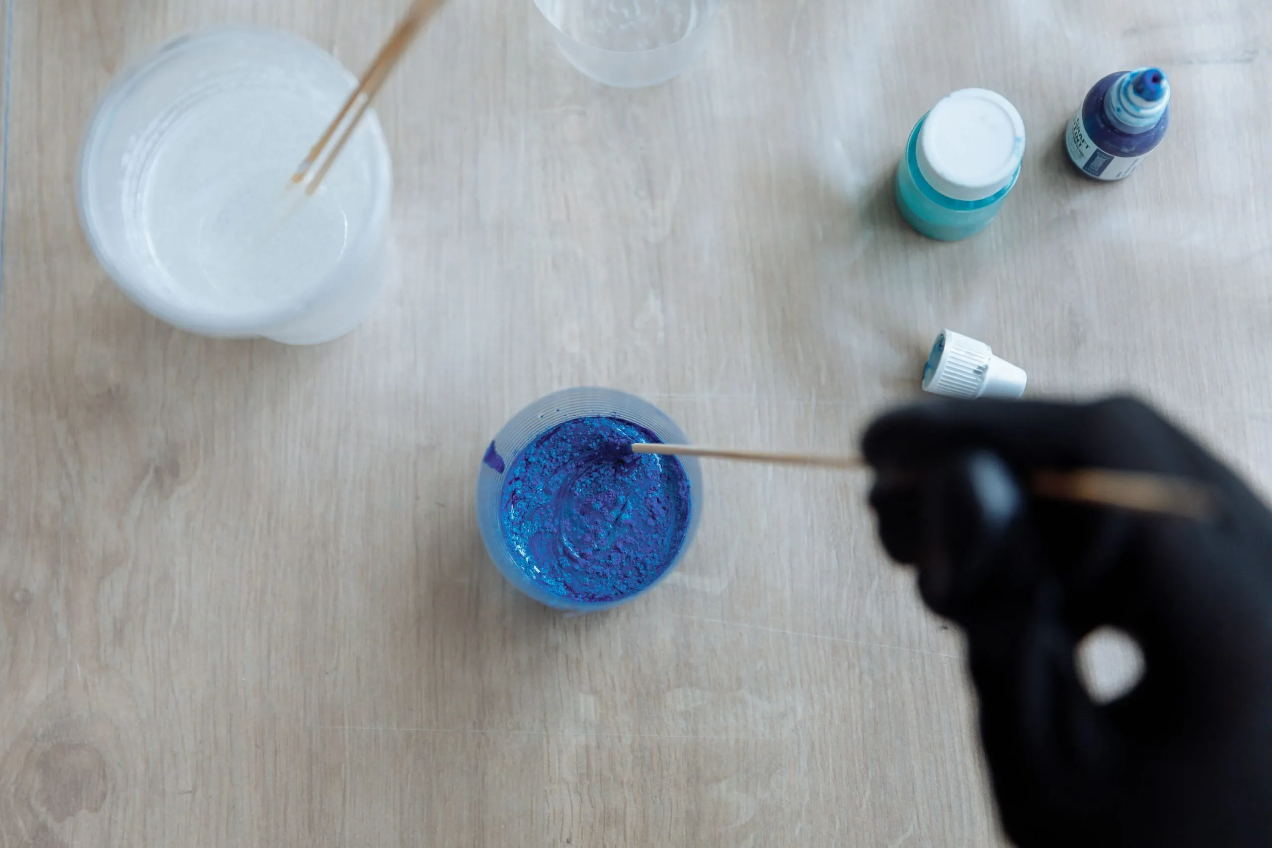 Close-up of hands mixing blue and white paints in clear cups with sticks on a wooden surface.