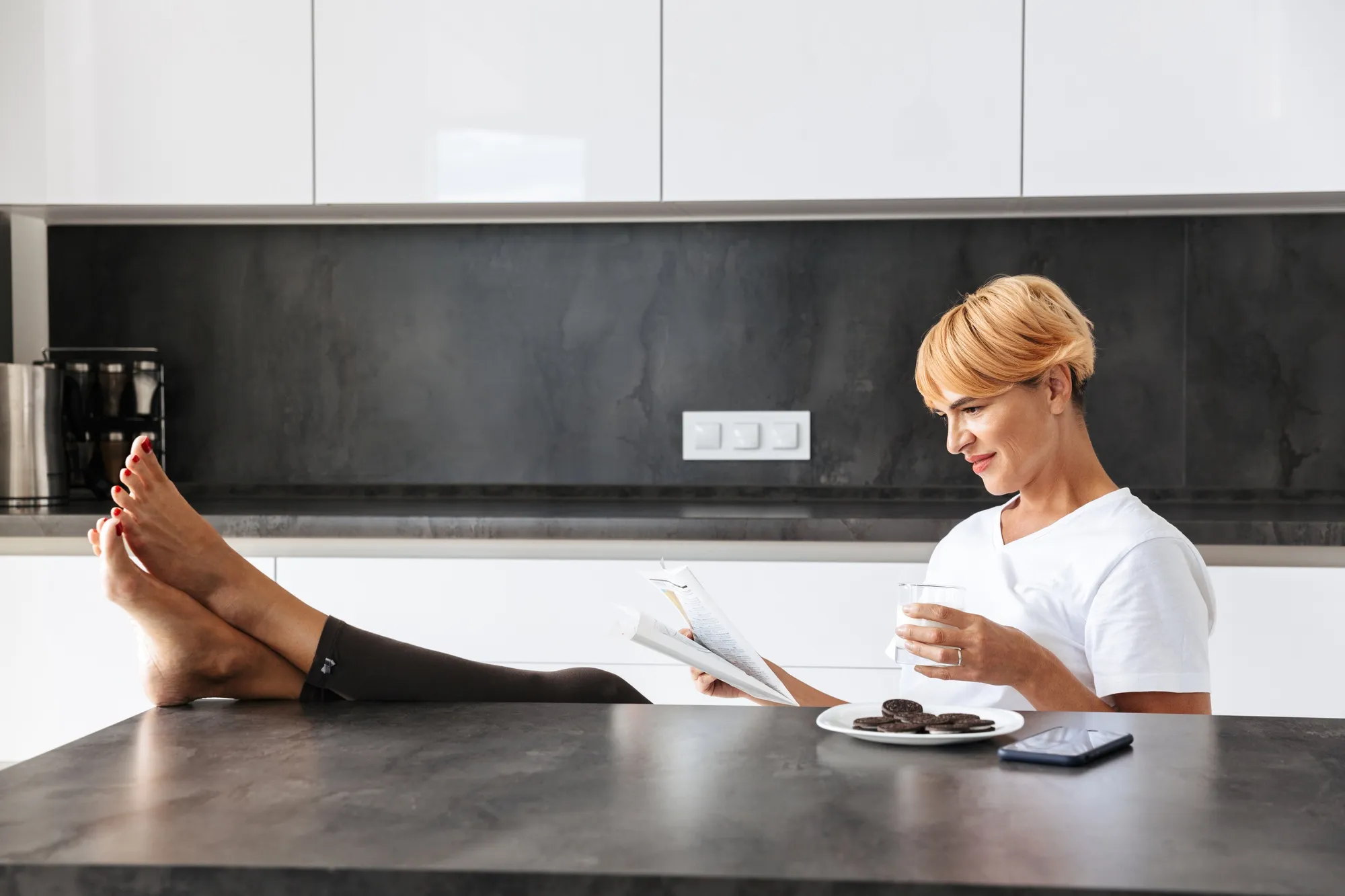 Pretty woman reading a newspaper while sitting at the kitchen table, drinking milk from a glass, eating cookies