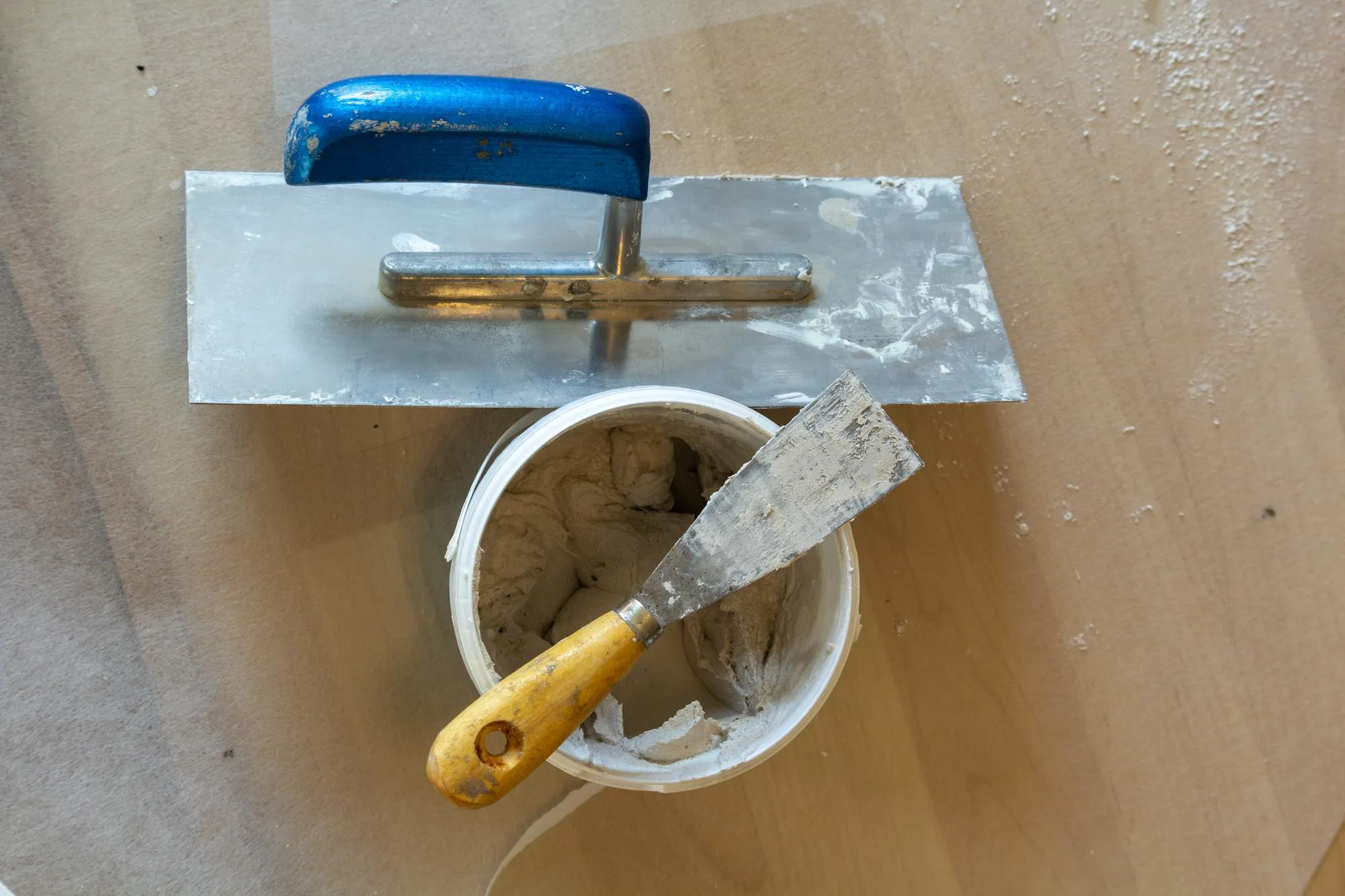 A small spatula on the bucket with fresh putty next to it a smoothing disc