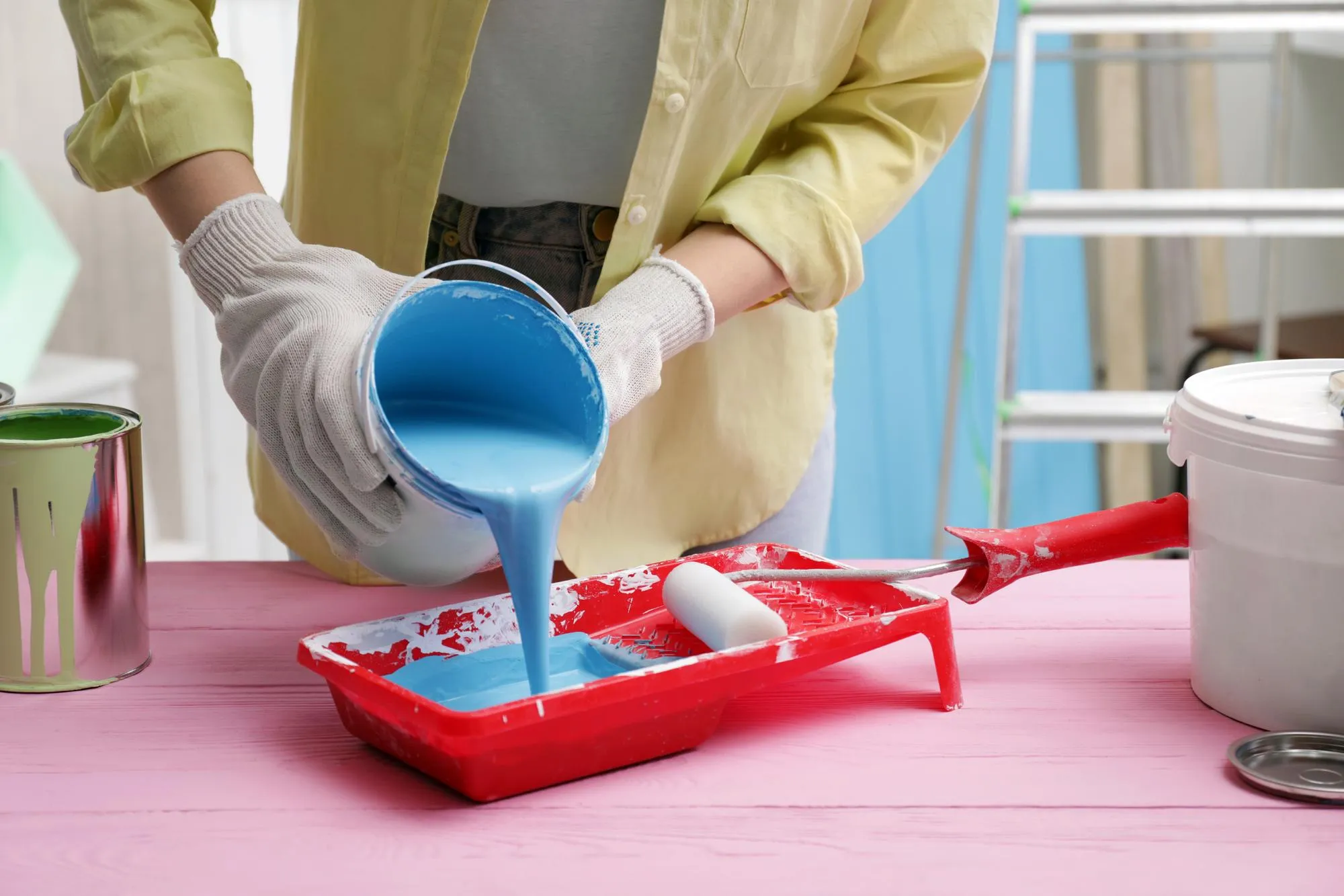Woman pouring light blue paint from can into tray at pink wooden