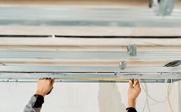 Worker's hands measure and adjust a metal profile for mounting a plasterboard ceiling frame, close-up, selective focus in the hands of a specialist. Industrial renovation and renovation