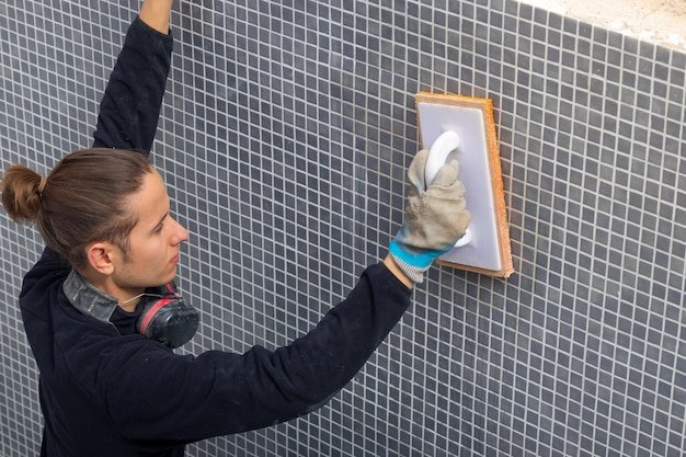 Young laborer man working on a swmming pool tiling with a plastering sponge