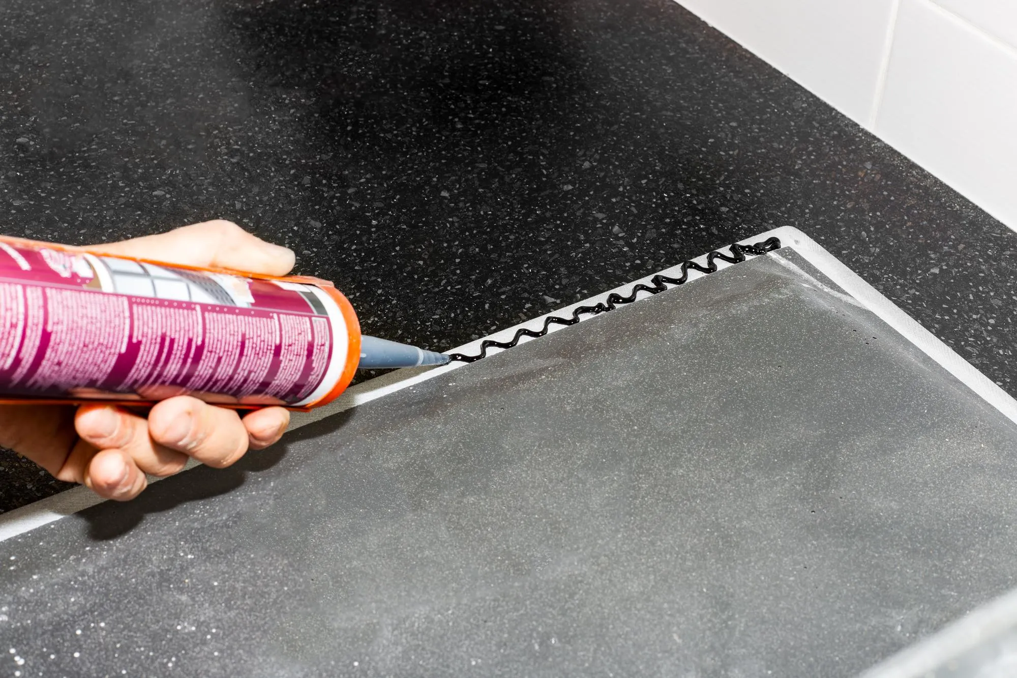 A man applies silicone caulk with a gun to the edge of a kitchen sink to install on the countertop