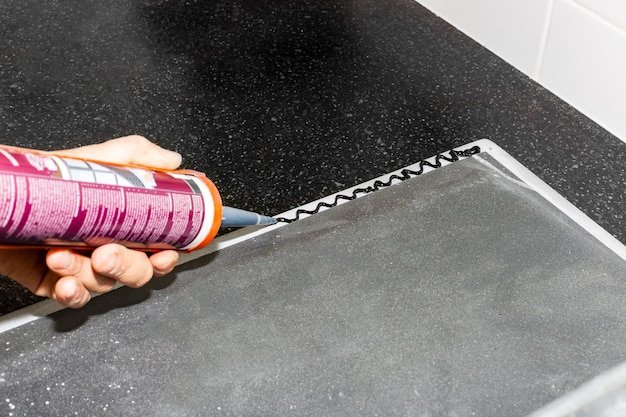 A man applies silicone caulk with a gun to the edge of a kitchen sink to install on the countertop
