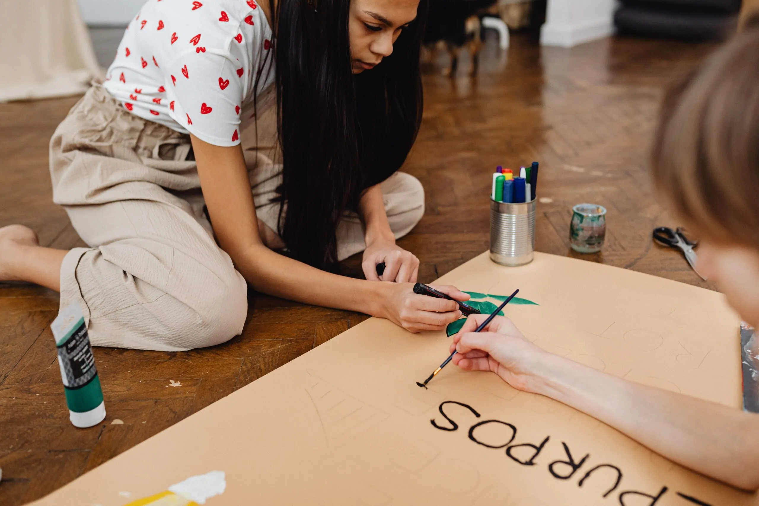 Two young adults engaged in a creative art project indoors, painting a poster with brushes and markers.
