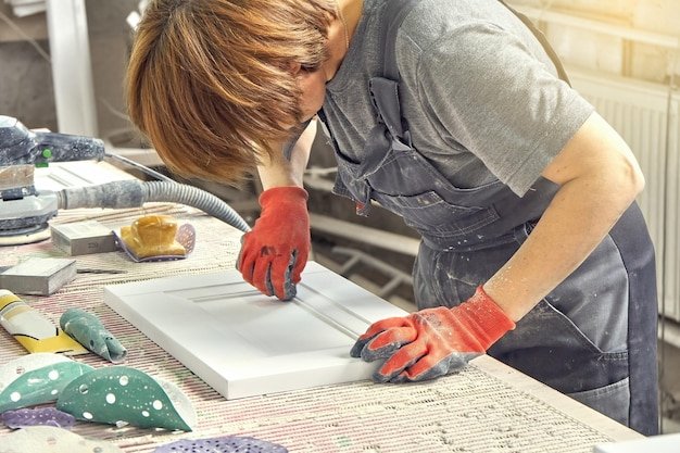 Female employee in uniform and working gloves polishes wooden part of furniture facade at workbench