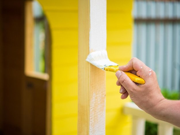 A man's hand with a brush paints a wooden beam with white paint