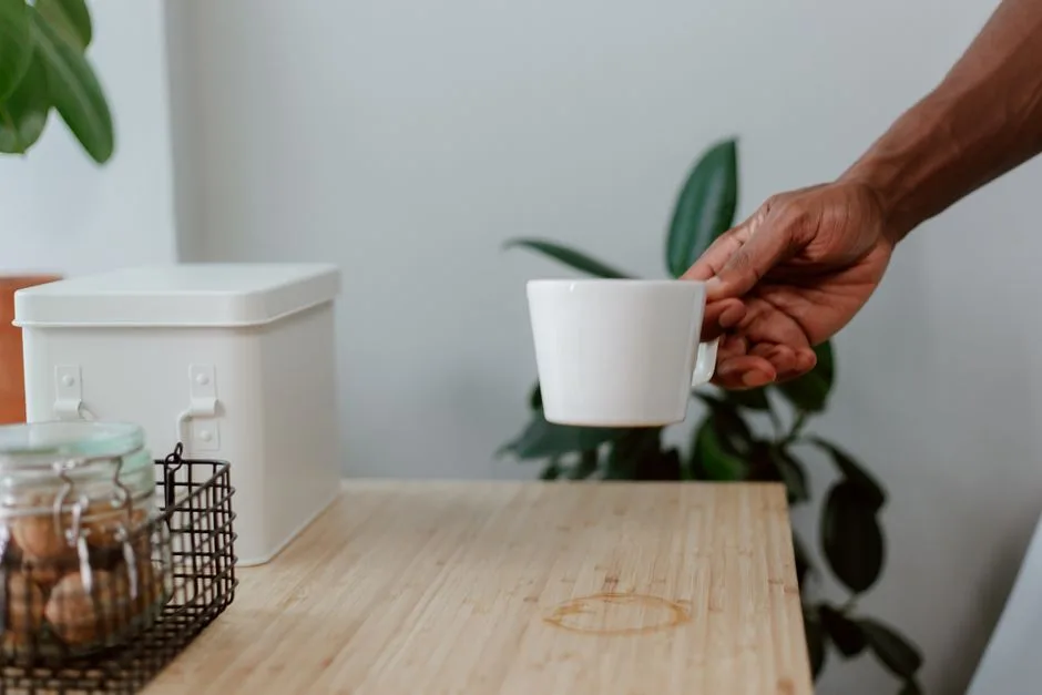 A hand holding a white coffee cup over a kitchen counter with a coffee stain and jars, tablescape setting.