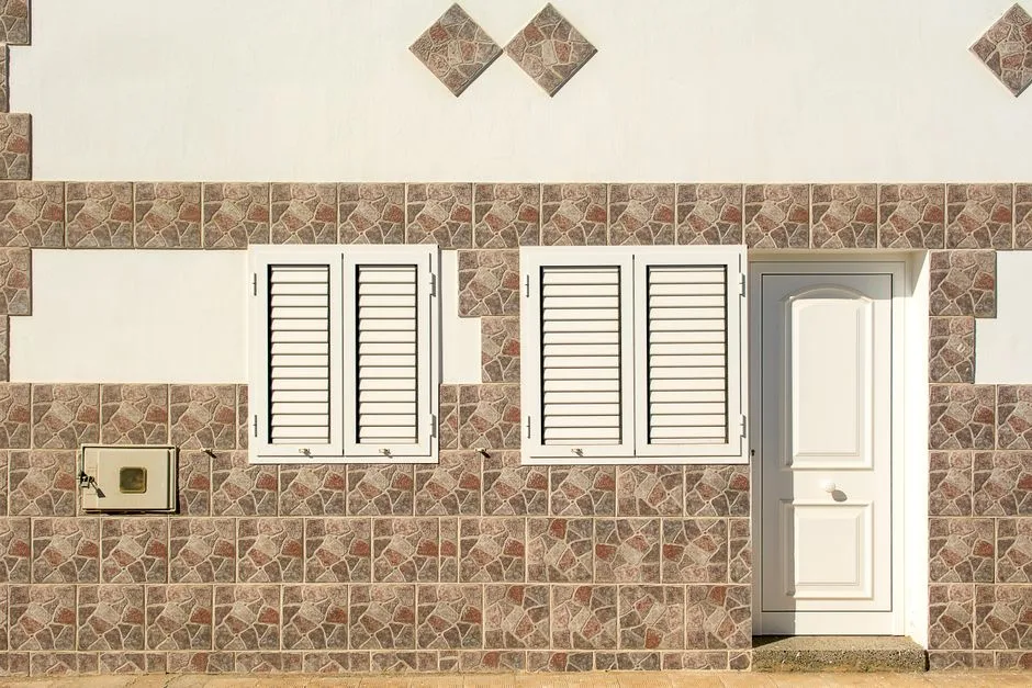 Close-up of a building facade featuring two white shuttered windows and a white door set within a mosaic tile wall.