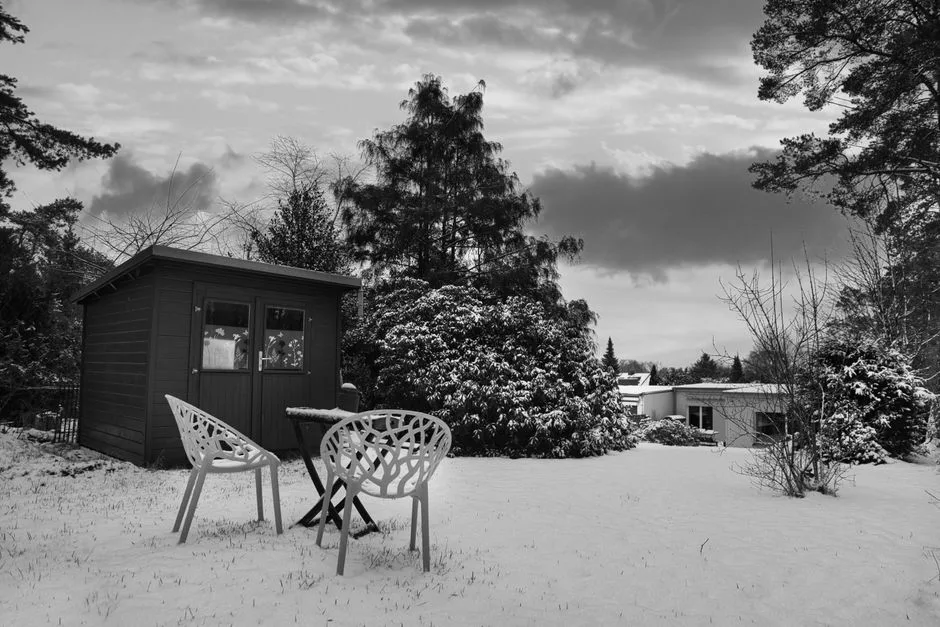 Black and white winter garden scene with snow-covered hut and chairs.
