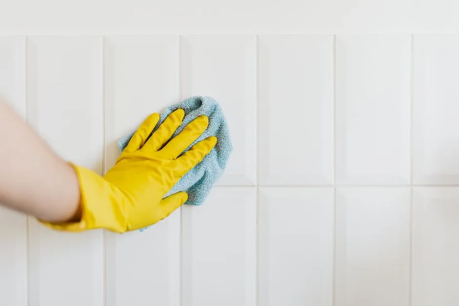 Crop anonymous housekeeper in yellow gloves cleaning white tiled wall using rag in bathroom
