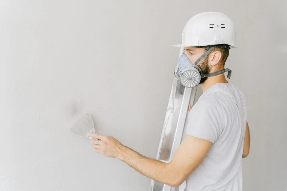 A construction worker wearing a gas mask and hard hat applies plaster to a wall indoors.