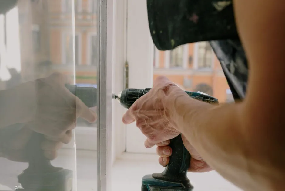 Unrecognizable professional male worker assembling white window with cordless screwdriver with drill near windowsill with reflection on glass during repair works in apartment