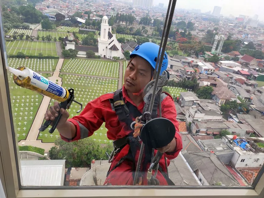 A professional window cleaner in Jakarta using safety gear and sealant with a cityscape vista backdrop.