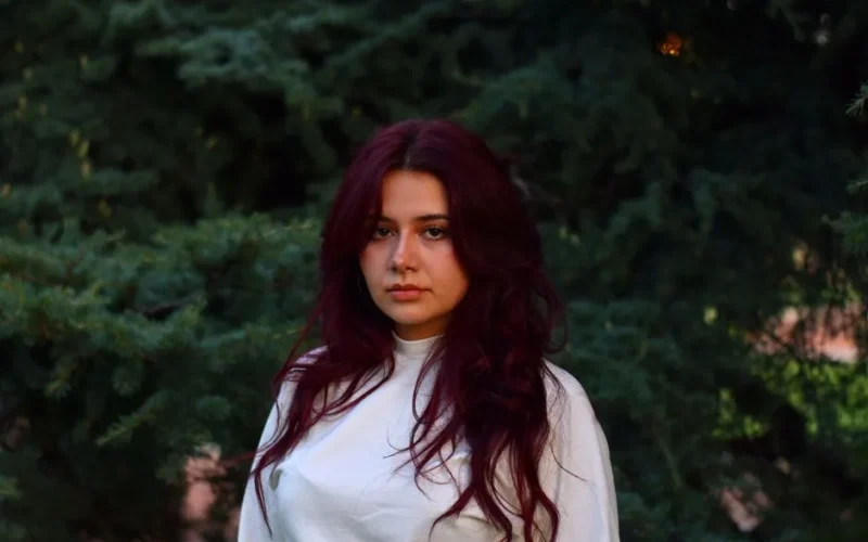 A woman with long burgundy hair poses outside in front of lush greenery in Mendoza, Argentina.