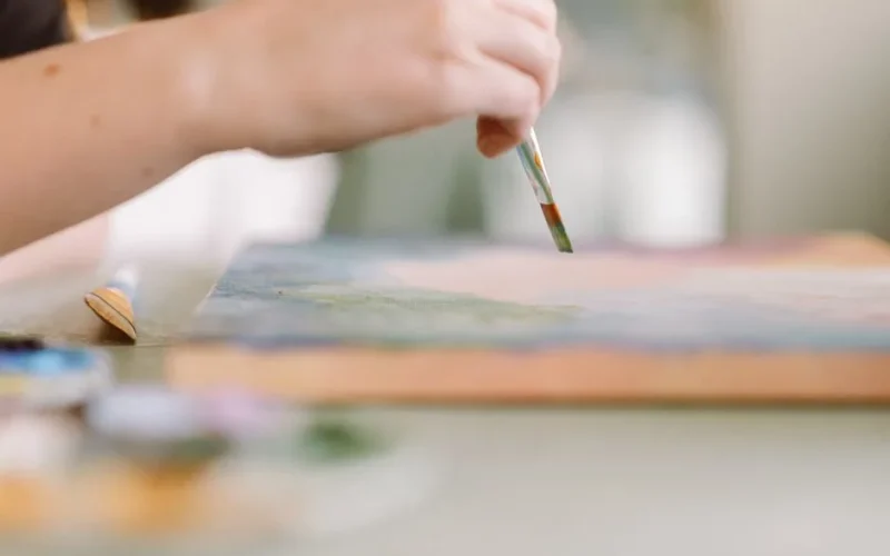 A female artist's hand skillfully painting on a canvas in a sunlit home studio setting.