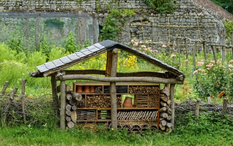 A handmade insect hotel in a lush rural garden, surrounded by vibrant flowers and rustic fencing.