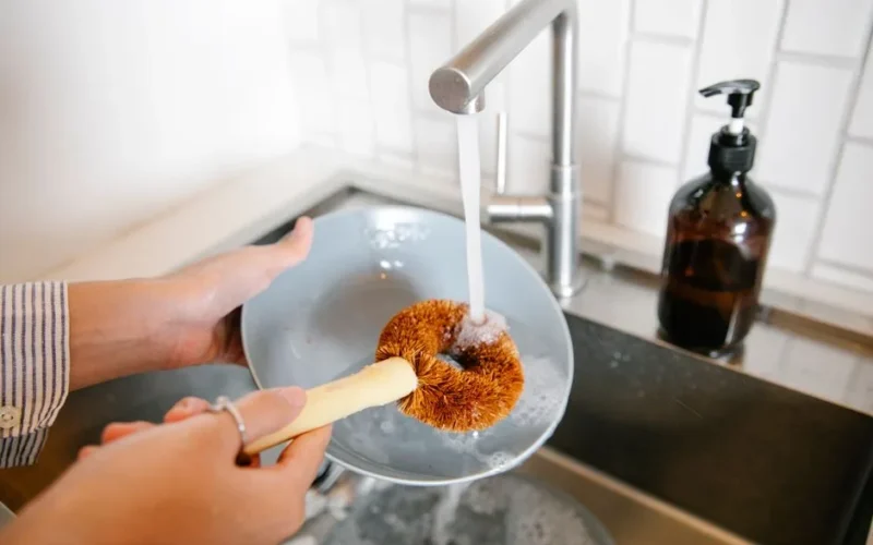 Unrecognizable female with brush washing plate at sink with pouring water and detergent while standing in light kitchen at home