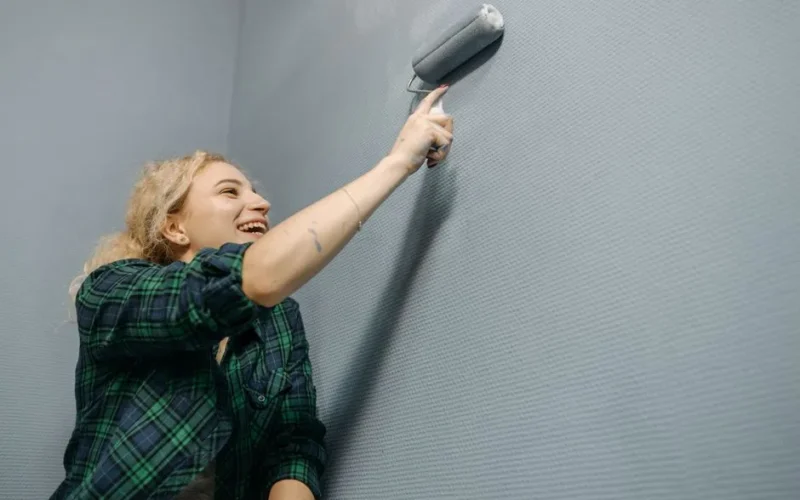 Laughing woman painting a blue wall indoors with a roller brush during home renovation.
