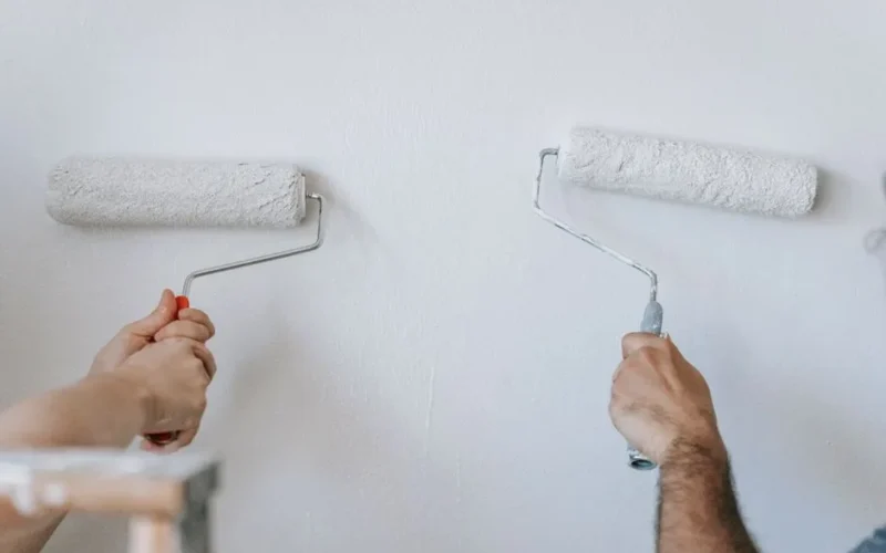 Close-up of two individuals painting a wall with paint rollers indoors.