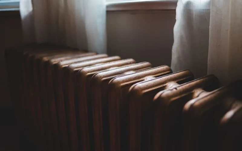 Aged heating battery made of cast iron near windowsill with curtains in house