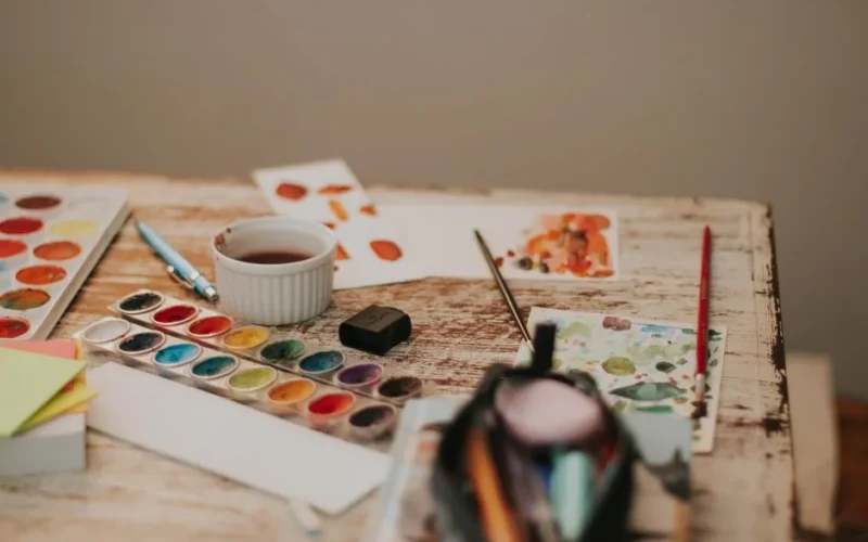 A cluttered artist's desk with watercolor paints, brushes, papers, and a cup, offering a glimpse into creative processes.