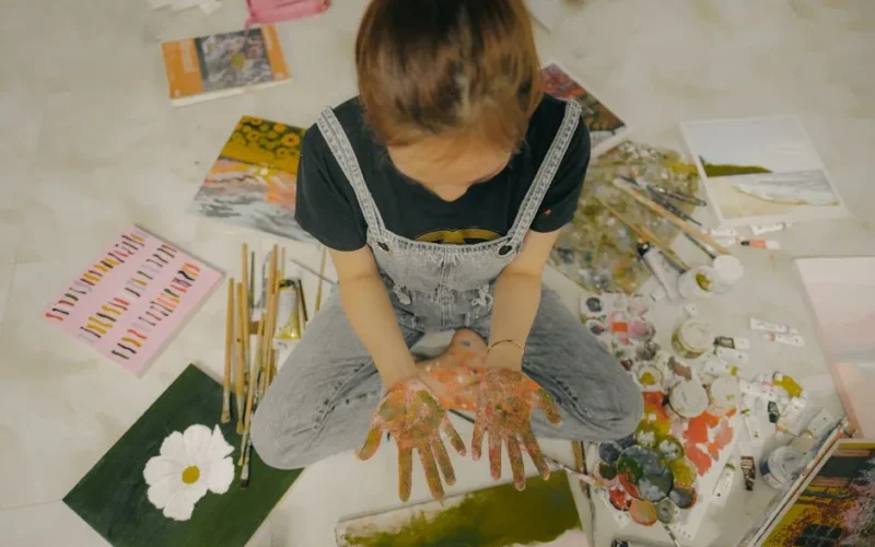 From above crop anonymous girl in casual jumpsuit demonstrating dirty hands while sitting on floor near paints paintbrushes and drawings