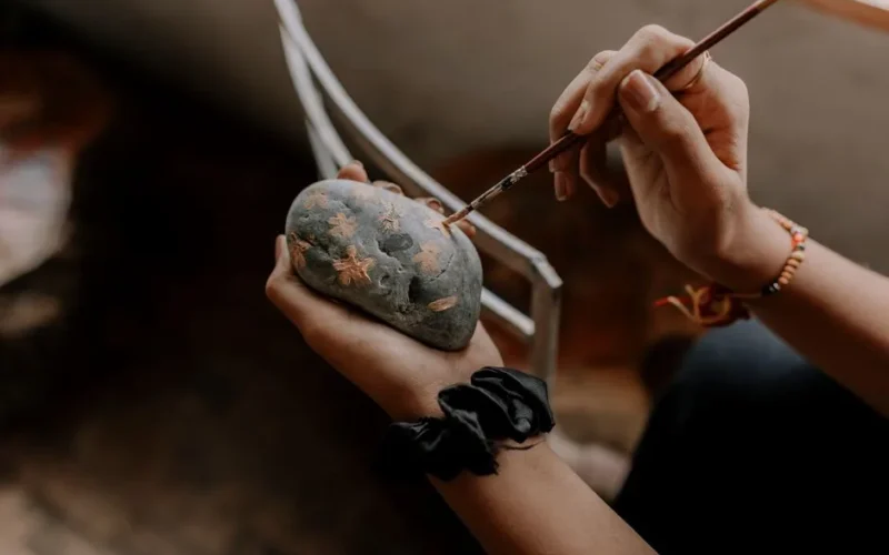 Close-up of hands painting intricate designs on a stone, showcasing artistic creativity indoors.