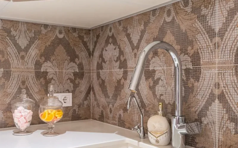 Contemporary kitchen stone counter with undercounter sink and shiny metal faucets at beige and brown tiled backsplash with ornament