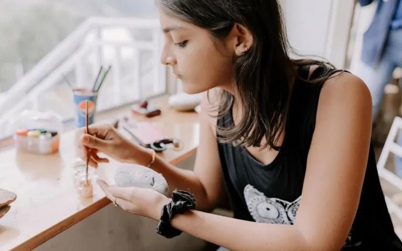 A young woman focused on painting stones by a sun-drenched window in an art studio.