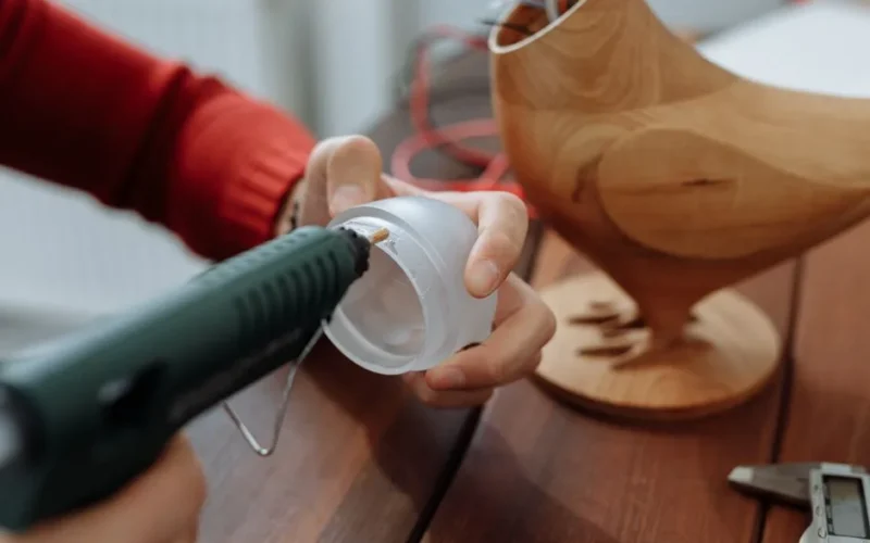 Close-up of a person gluing a plastic lid next to a wooden chicken model on a table.