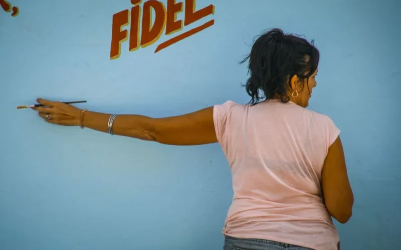 A woman artist paints red letters on a light blue wall, showcasing creative expression.