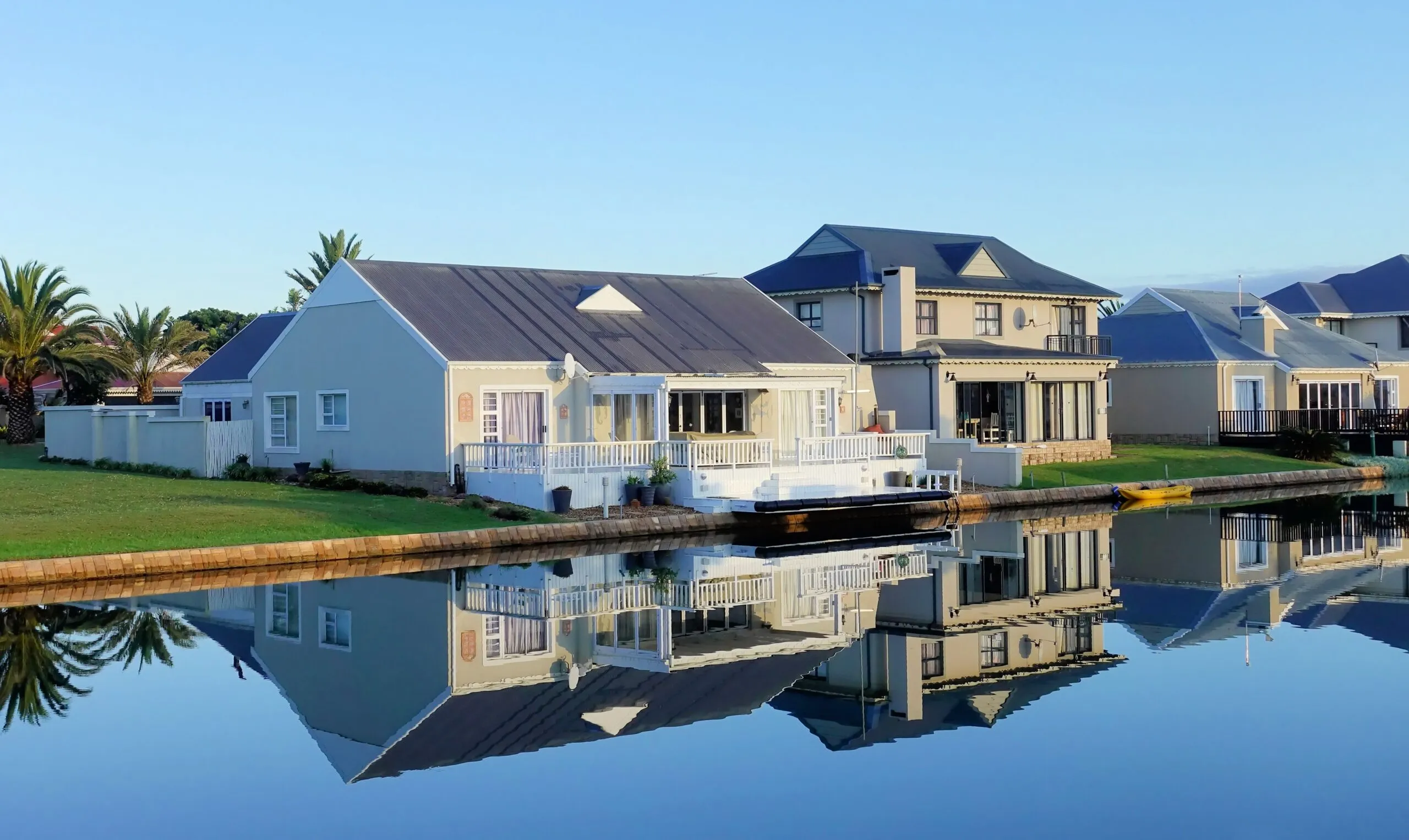 Luxury modern homes reflecting on a peaceful, clear lake during daylight, showcasing architectural beauty.