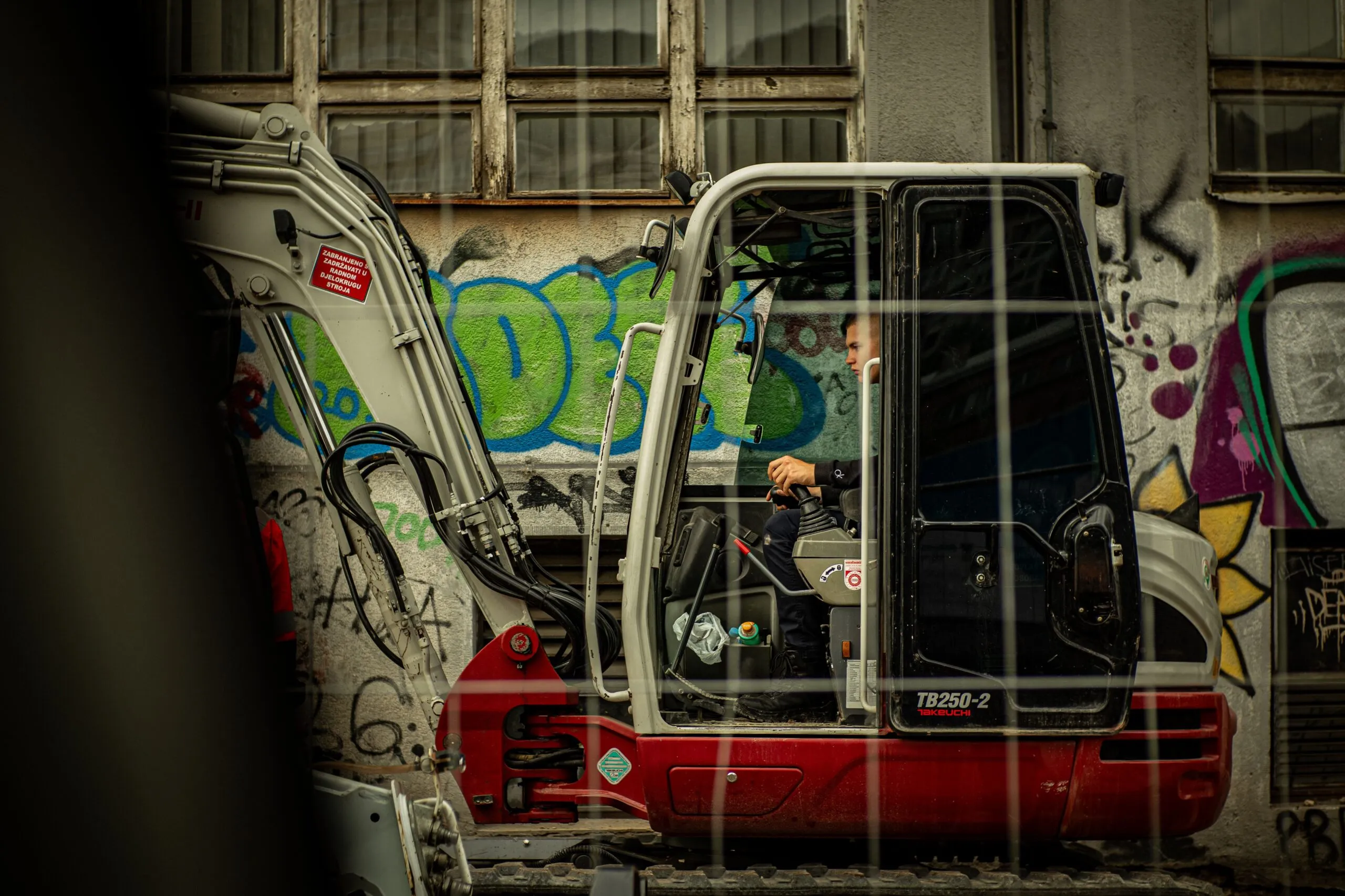 Man operating construction digger in urban setting with graffiti background.