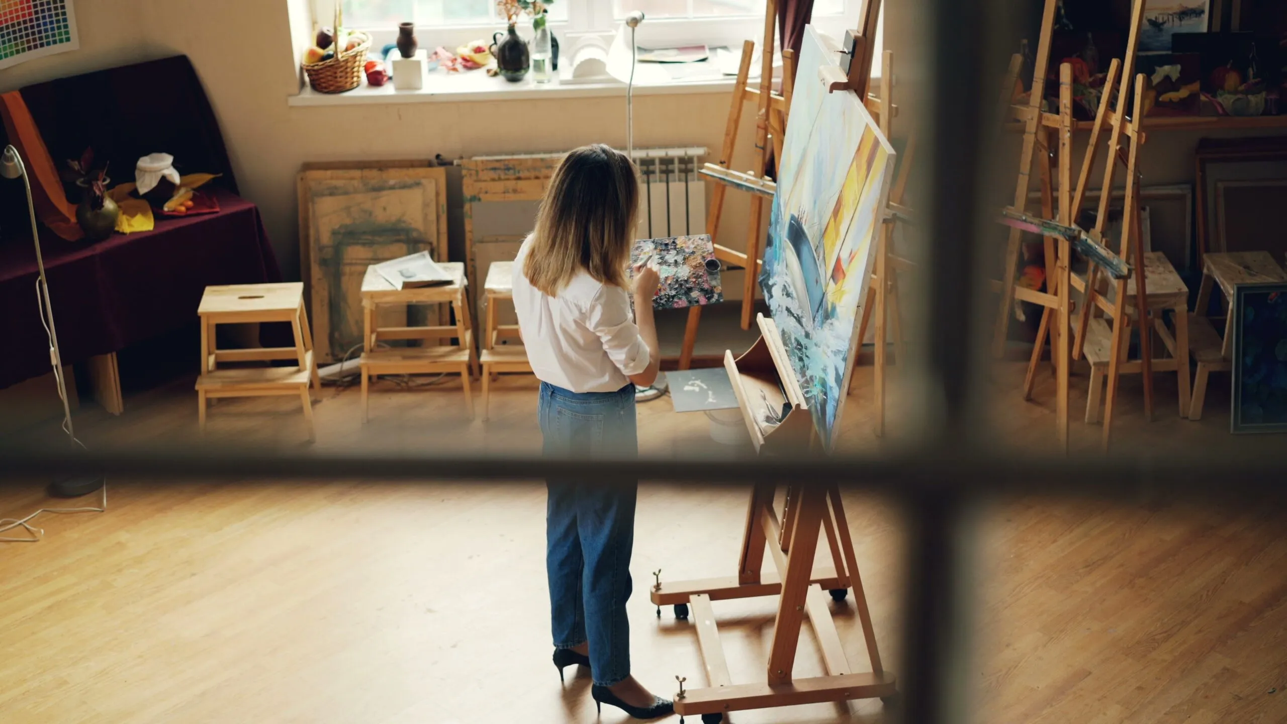 Artist focused on painting in a well-lit studio filled with art supplies.