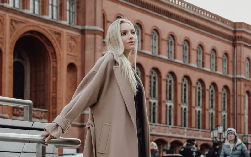 Stylish woman with long blonde hair poses elegantly in front of a historic red brick building in an urban setting.