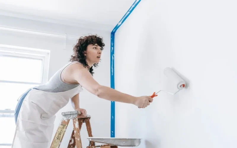 A woman stands on a ladder painting a wall white, part of a DIY home renovation.