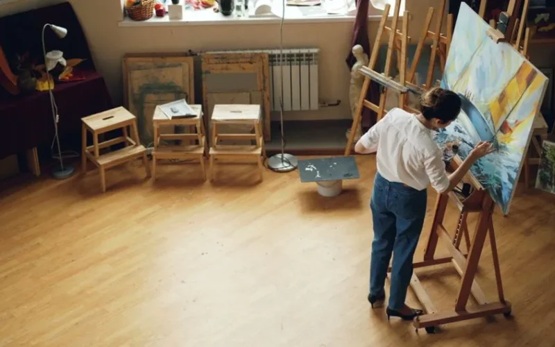 A woman painting on a canvas in a well-lit artist studio filled with creative supplies.