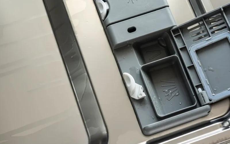 Closeup of an open dishwasher detergent compartment with water droplets visible.