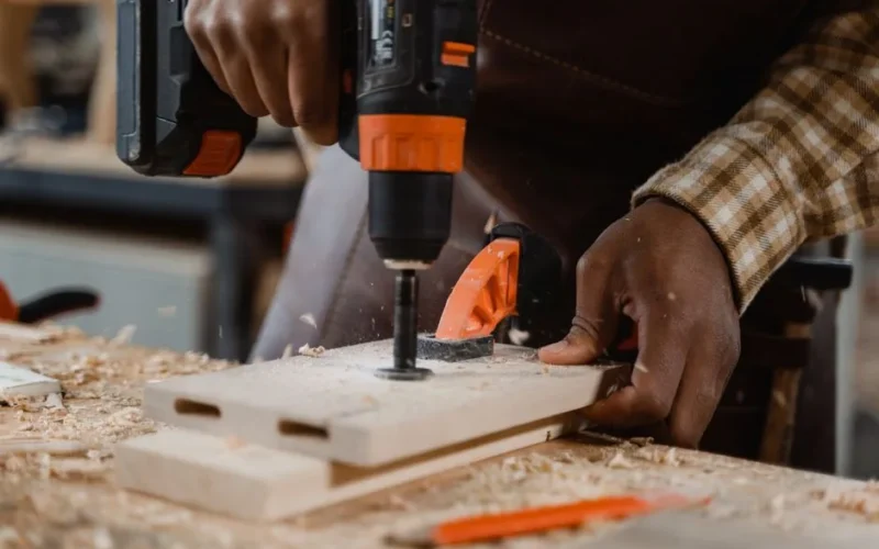 Carpenter uses drill on wood board, surrounded by sawdust in a workshop setting.