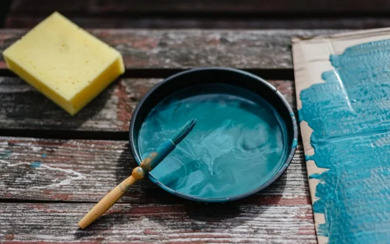 From above of turquoise paint in bowl with brush placed on wooden board near cardboard and sponge during repair work